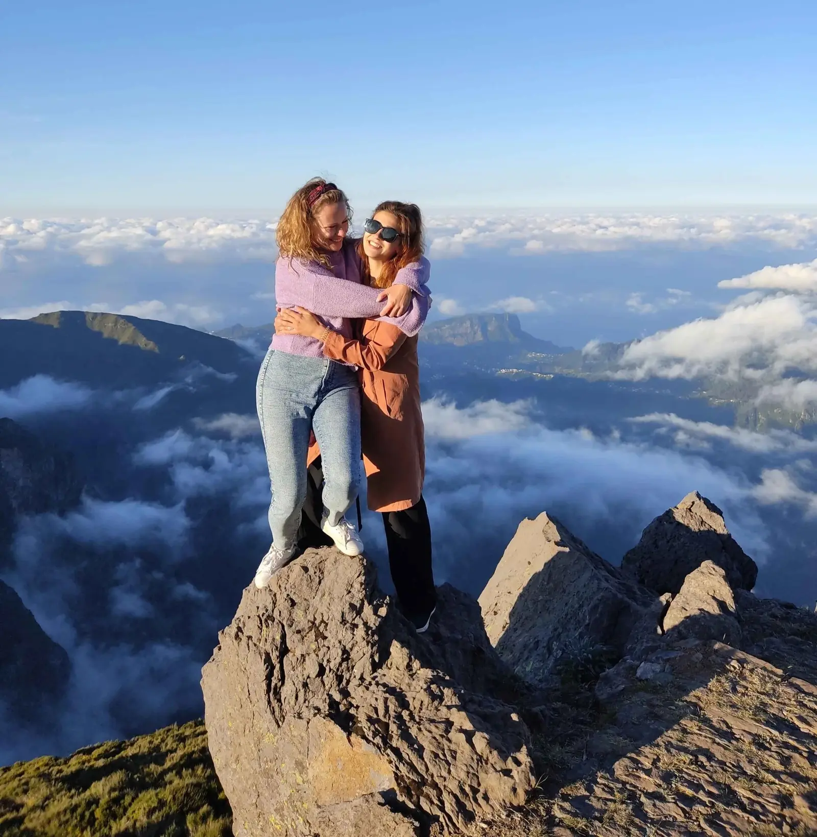 Two friends hugging on a rocky peak above a sea of clouds
